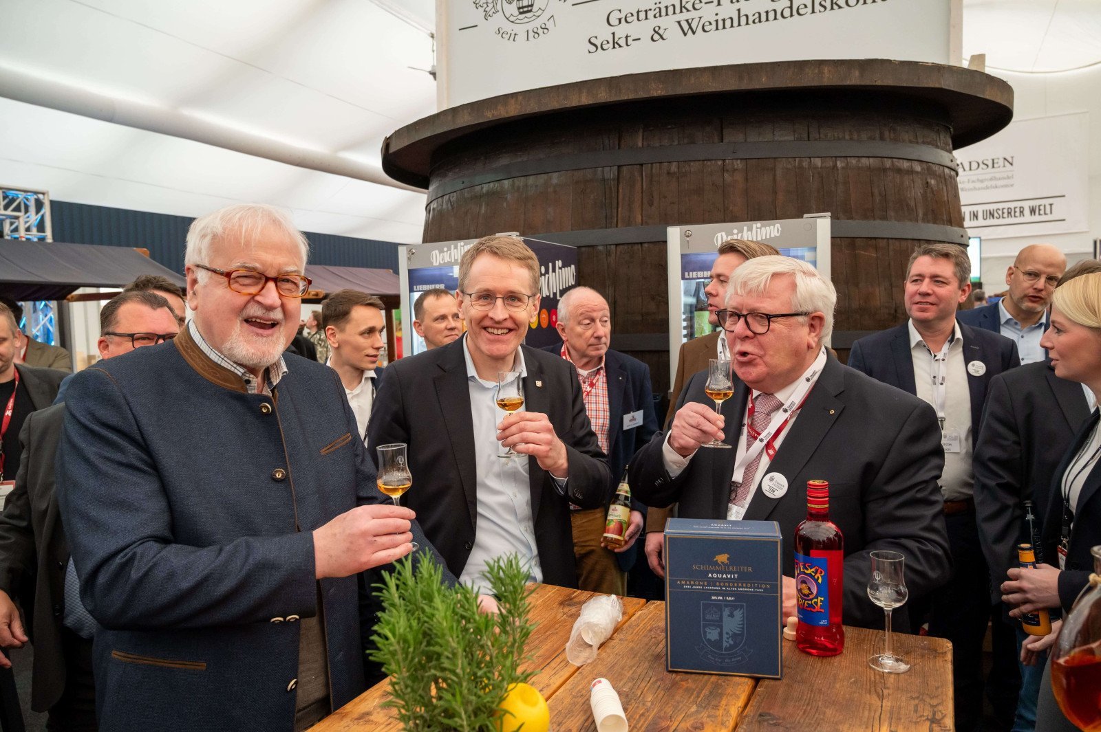 Mehrere Personen stehen an einem Holztisch und halten kleine Gl&auml;ser mit Getr&auml;nken in der Hand, im Hintergrund ein gro&szlig;es rundes Holzfass und ein Schild mit Text.